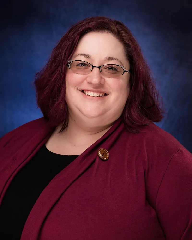 Corporate headshot of a woman in a dark red blazer photographed on a blue mottled studio backdrop